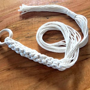 May include: A white macrame plant hanger with a woven section and a wooden ring. The long, twisted cotton cords are arranged in a spiral, with a section of loose cords at the top. The plant hanger is displayed on a wooden surface.