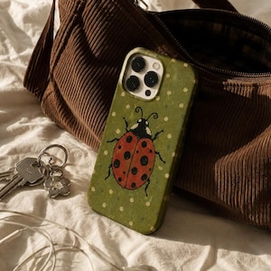 May include: A green smartphone case featuring a red ladybug with black spots and antennae, set against a background of small white dots. The case is positioned next to a brown corduroy shoulder bag, a metal keyring with a flower charm, and white wired earbuds.