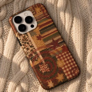 May include: A smartphone case featuring a patchwork design with autumn-themed patterns including red and white gingham, maple leaves, and textured brown and green elements. The case has a cutout for a triple-lens camera system on the back.