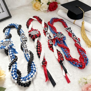 May include: Braided graduation leis in red, white, blue, and black. Each lei features a name and "Class 2026" ribbon. A graduation cap and diploma are in the background, along with flowers.
