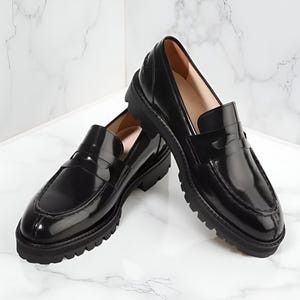 Pode incluir: Um par de mocassins de couro preto com acabamento brilhante. Os sapatos têm um design clássico de mocassim com sola grossa e com ranhuras. Os sapatos são exibidos em uma superfície de mármore branco.
