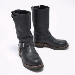 Puede incluir: Un par de botas de moto de cuero negro. Las botas son de media caña, con correas con hebilla y suelas marrones. El diseño es clásico y robusto. Miden aproximadamente 30 cm de altura.