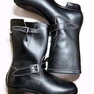 Puede incluir: Un par de botas de cuero negro. Las botas son de media caña, con punta redonda, tacón bajo y correas ajustables con hebilla. Diseño clásico y robusto, adecuado para varios conjuntos.