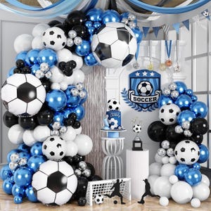 May include: A soccer-themed party decoration set with a balloon arch, soccer ball balloons, and a cake. The color scheme includes white, black, and blue. A soccer trophy and a goal are also present.