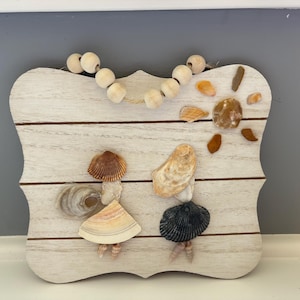 Puede incluir: Placa decorativa de madera blanqueada, adornada con conchas marinas y cuentas de madera. El diseño presenta dos figuras hechas de conchas, un sol y una guirnalda de cuentas. La placa es una pieza única de decoración costera.