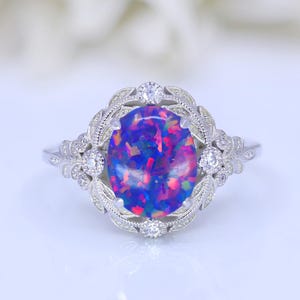 May include: A silver ring with an oval opal gemstone, displaying blue, purple, and pink colours. The ring has an ornate setting with small accent stones and a leaf design. The gemstone's dimensions are approximately 1.5 cm x 1 cm.