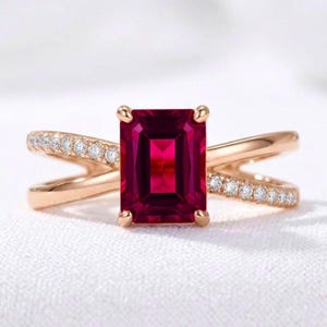 Peut inclure: Une bague en or rose ornée d'une grande pierre précieuse rouge foncé taille émeraude au centre. L'anneau présente un design croisé avec un côté agrémenté d'une rangée de petites pierres blanches rondes, tandis que l'autre côté est lisse.