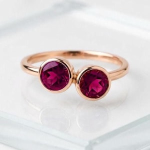 May include: A rose gold ring featuring two round, bezel-set deep pink gemstones. The band is smooth and polished, with the two stones positioned side-by-side in a bypass style setting, creating a balanced and symmetrical appearance on the finger.