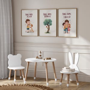 Puede incluir: Tres impresiones artísticas enmarcadas con las frases "YOU ARE KIND", "YOU ARE BRAVE" y "YOU ARE LOVED". Una mesa blanca y dos sillas, una con forma de oso y otra con forma de conejo, están frente a las impresiones. Una pequeña faro de madera y bloques están sobre la mesa.