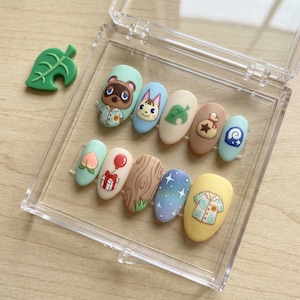 Puede incluir: Un conjunto de diez u&ntilde;as postizas con varios dise&ntilde;os, incluyendo personajes de dibujos animados, una hoja, un regalo y una camisa. Las u&ntilde;as se muestran en una caja de pl&aacute;stico transparente, con un dije verde en forma de hoja.