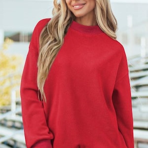 May include: A bright red long-sleeved sweatshirt featuring a mock neck collar, dropped shoulders, and ribbed cuffs at the wrists and hem. The garment is constructed from a textured fleece material and is paired with light blue denim shorts.