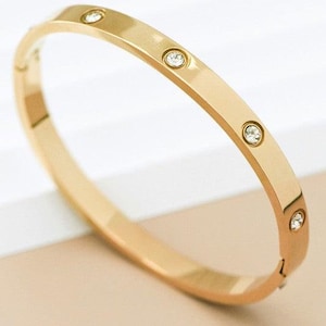 May include: A gold-colored bangle bracelet with five clear, round gemstones. The bracelet has a smooth, polished surface and a simple, elegant design. The gemstones are evenly spaced along the top of the bracelet.