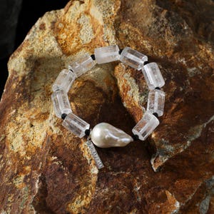 May include: A bracelet featuring clear, rectangular-cut beads strung together with black cord. A large, irregular-shaped pearl and a small silver tag with the text "PURAPERIA" are also included in the design. The jewelry is displayed on a textured, brown rock surface.