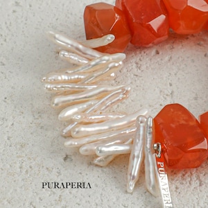 Puede incluir: Primer plano de un collar con cuentas de piedras preciosas naranjas y detalles de perlas blancas alargadas. El collar tiene un cierre plateado con el nombre de la marca "PURAPERIA" grabado. El collar se muestra sobre una superficie clara y texturizada.