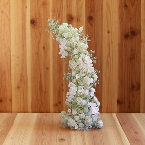 以下が含まれることがあります： 結婚式などに使われる、白い花と緑の垂直方向のアレンジメント。 花は密集しており、曲線を描いています。 背景は木製の壁と床です。