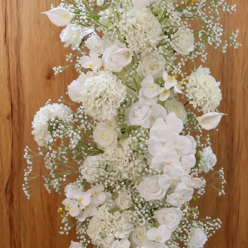 以下が含まれることがあります： 木製の背景に、バラ、アジサイ、蘭、かすみ草などの白い花が垂直に配置されています。 花のディスプレイは密度が高く、さまざまな質感と形があり、視覚的に魅力的な構成になっています。