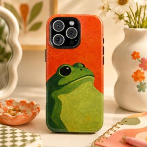 May include: A smartphone case featuring an oil painting style illustration of a green frog against a vibrant orange background. The case has a cutout for a triple-lens camera system and is designed to fit a modern smartphone with a black camera module.