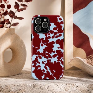 May include: A smartphone case featuring a high-contrast, irregular cow-print pattern in deep maroon and light baby blue. The case has a precise cutout for the rear camera module, which contains three circular lenses and a flash component.