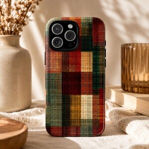 May include: A rectangular phone case featuring a multi-colored plaid pattern with vertical and horizontal lines in shades of deep red, forest green, mustard yellow, and dark brown, with a black camera cutout area at the top left corner.