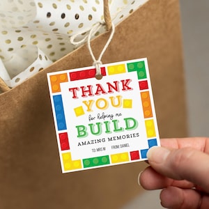 Pode incluir: Uma etiqueta de presente colorida com as palavras "THANK YOU" e "BUILD AMAZING MEMORIES" em uma fonte lúdica. A etiqueta é projetada para se assemelhar a blocos de construção de brinquedo, com detalhes em vermelho, amarelo, azul e verde. A etiqueta está presa a uma sacola de presente marrom.