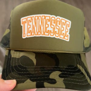 May include: An olive green and camouflage trucker hat featuring the word "TENNESSEE" in orange block letters. The hat has a mesh back and a curved peak. The hat is being held by a person.