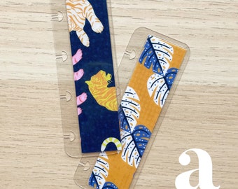 Può includere: Due segnalibri decorativi con custodie in plastica trasparente. Uno presenta uno sfondo blu navy con illustrazioni di tigri arancioni e zampe rosa. L'altro ha uno sfondo arancione con disegni di foglie blu e bianche.