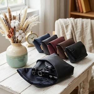 May include: A collection of five small, rectangular, flap-closure pouches in navy, charcoal, burgundy, brown, and black. One black pouch is open, revealing a black camera inside. The pouches are arranged on a white wooden surface near a vase.