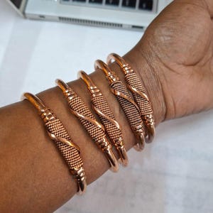 Op de afbeelding: Een verzameling koperkleurige metalen armbanden. Elke armband heeft een gladde, afgeronde band met een getextureerd, geweven ontwerp in het midden. De armbanden worden om een pols gedragen en tonen hun warme, metallic tint en ingewikkelde details.