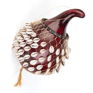 May include: A dark red, polished gourd percussion instrument, known as a shekere, featuring a net of white cowrie shells covering the rounded body. The instrument has a curved neck and a small, light brown fiber tassel hanging from the base.