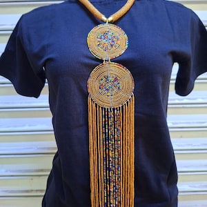 May include: A gold beaded necklace with two large circular pendants and a long fringe. The pendants feature concentric circles of gold beads with colorful accents. The necklace is displayed on a dark blue shirt.