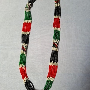Puede incluir: Un collar de cuentas con secciones alternas de cuentas rojas, negras, verdes y blancas. El collar está hecho de múltiples hilos de pequeñas cuentas de colores, creando un diseño audaz y llamativo.
