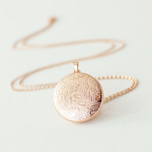 May include: A rose gold locket necklace with a delicate chain. The round locket features an intricate floral pattern. The necklace is displayed on a white surface, with the chain gently curving. The locket is a classic jewelry piece.