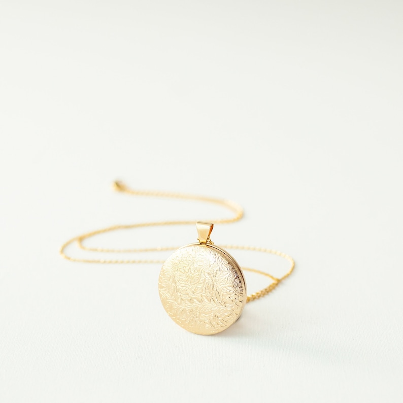 Peut inclure: Un pendentif m&eacute;daillon circulaire dor&eacute; orn&eacute; d'un motif floral en relief complexe sur sa face avant. Le m&eacute;daillon est fix&eacute; &agrave; une fine cha&icirc;ne dor&eacute;e assortie qui s'&eacute;tend vers l'ext&eacute;rieur, pr&eacute;sentant une finition m&eacute;tallique polie.
