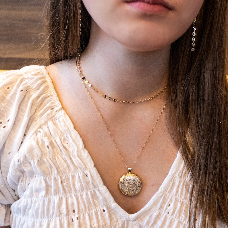 Peut inclure: Un ensemble de colliers superpos&eacute;s dor&eacute;s compos&eacute; d'une cha&icirc;ne courte &agrave; petits maillons circulaires plats et d'une cha&icirc;ne plus longue avec un pendentif rond textur&eacute;. Le pendentif pr&eacute;sente un motif en relief d&eacute;taill&eacute;. Port&eacute; avec un chemisier blanc.