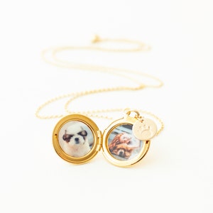 May include: A gold locket necklace with two small dog photos inside. The locket is open, showing the pictures. A small charm with a paw print hangs next to the locket. The necklace is set against a white background.