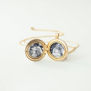 Peut inclure: Un pendentif m&eacute;daillon circulaire dor&eacute; sur une fine cha&icirc;ne assortie. Le m&eacute;daillon est ouvert, r&eacute;v&eacute;lant deux photographies circulaires en noir et blanc &agrave; l'int&eacute;rieur. Une photo montre un adulte tenant un b&eacute;b&eacute;, l'autre un adulte souriant avec un b&eacute;b&eacute;.