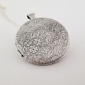 Peut inclure: Un m&eacute;daillon rond en argent orn&eacute; d'un motif en relief complexe repr&eacute;sentant de petites fleurs et des feuilles d&eacute;taill&eacute;es sur toute la surface. Le m&eacute;tal pr&eacute;sente une finition sombre dans les zones en creux pour souligner le motif floral.