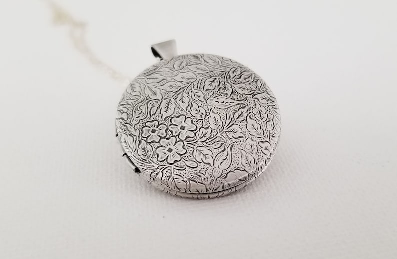 Peut inclure: Un m&eacute;daillon rond en argent orn&eacute; d'un motif en relief complexe repr&eacute;sentant de petites fleurs et des feuilles d&eacute;taill&eacute;es sur toute la surface. Le m&eacute;tal pr&eacute;sente une finition sombre dans les zones en creux pour souligner le motif floral.