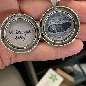 Peut inclure: Un m&eacute;daillon rond en m&eacute;tal argent&eacute; ouvert r&eacute;v&eacute;lant deux compartiments circulaires. Le c&ocirc;t&eacute; gauche contient un papier blanc avec le texte manuscrit "I love you, baby" &agrave; l'encre noire. Le c&ocirc;t&eacute; droit affiche une image &eacute;chographique en noir et blanc.