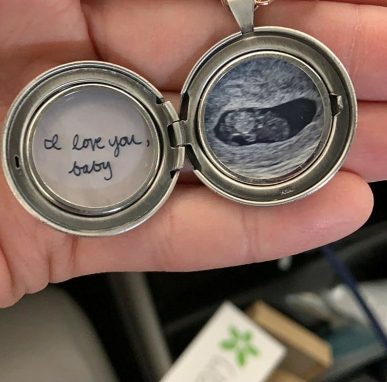 Peut inclure: Un m&eacute;daillon rond en m&eacute;tal argent&eacute; ouvert r&eacute;v&eacute;lant deux compartiments circulaires. Le c&ocirc;t&eacute; gauche contient un papier blanc avec le texte manuscrit "I love you, baby" &agrave; l'encre noire. Le c&ocirc;t&eacute; droit affiche une image &eacute;chographique en noir et blanc.