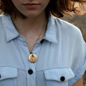 Peut inclure: Un m&eacute;daillon rond en or orn&eacute; d'une gravure florale complexe et d'un petit d&eacute;tail en forme de c&oelig;ur, suspendu &agrave; une fine cha&icirc;ne dor&eacute;e. Le m&eacute;daillon est port&eacute; sur une chemise boutonn&eacute;e bleu clair avec deux poches poitrine et des boutons fonc&eacute;s.