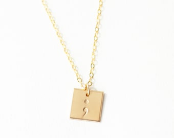 May include: A gold-coloured necklace with a square pendant featuring a semicolon symbol. The pendant is suspended from a delicate chain. The necklace is displayed against a white background.