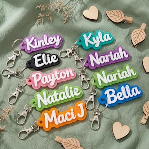 Op de afbeelding: Een verzameling gepersonaliseerde sleutelhangers met namen zoals Kinley, Kyla en Bella, in kleuren als paars, groen en oranje. De sleutelhangers liggen op een groene stoffen ondergrond, versierd met houten hartjes en blaadjes.