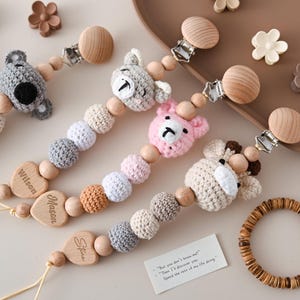 May include: A collection of baby pacifier clips with crochet animal heads, wooden beads, and personalized wooden heart-shaped name tags. The clips feature a koala, bear, pink bear, and giraffe. A wooden bracelet and decorative flowers are also present.