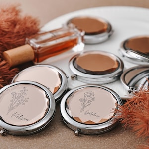 以下が含まれることがあります： 花柄と名前が刻まれたシルバーのコンパクトミラーのコレクションと、小さな香水瓶。 ミラーは薄いピンク色の背景で、白いお皿の上に配置されています。