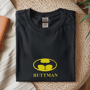 May include: Black t-shirt with a yellow graphic resembling the Batman logo, but with a butt shape. The word "BUTTMAN" is printed below the graphic in yellow. The shirt is folded and placed on a woven surface.
