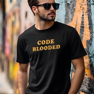 May include: Black crew neck t-shirt featuring the words "CODE BLOODED" in a yellow, distressed font. The background is a colourful graffiti wall. The t-shirt is a casual style.