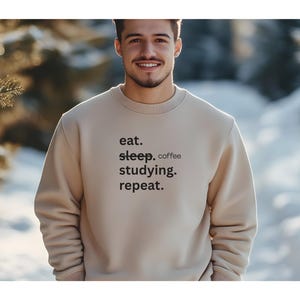 以下が含まれることがあります： ベージュのクルーネックのスウェットシャツ。黒で「eat. sleep. coffee. studying. repeat.」とプリントされています。スウェットシャツは柔らかい素材でできており、カジュアルウェアに適しています。背景はぼやけた屋外のシーンです。