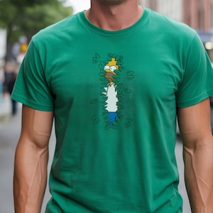 May include: Green t-shirt featuring a cartoon illustration of Homer Simpson peeking through leaves. The design includes Homer's face, white shirt, and blue pants. The shirt is short-sleeved and made of a soft material.