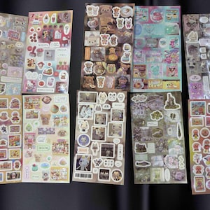 May include: A collection of sticker sheets featuring various cartoon characters, food items, and decorative elements. The stickers are in a variety of colors, including pink, blue, yellow, and brown. The sheets are rectangular and arranged on a dark surface.